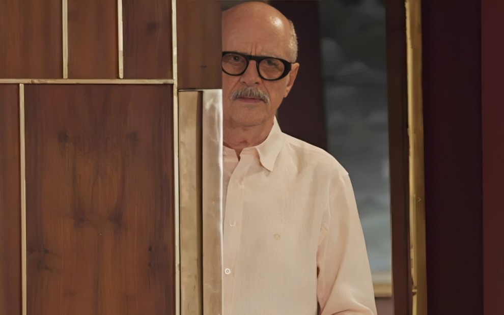 O ator Marcos Caruso com expressão séria em cena de Elas por Elas, atrás de porta
