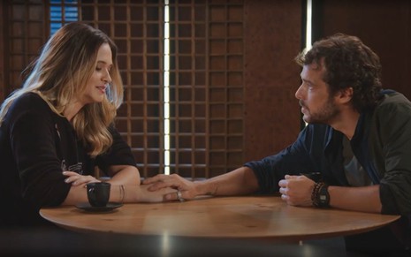 Em cena de Família É Tudo, Juliana Paiva e Jayme Matarazzo estão conversando em uma mesa e ele segura a mão dela
