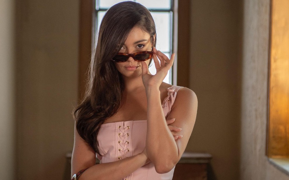 Sophie Charlotte usa blusa de alça rosa e segura um óculos escuro no rosto; ela está caracterizada como sua personagem em Renascer