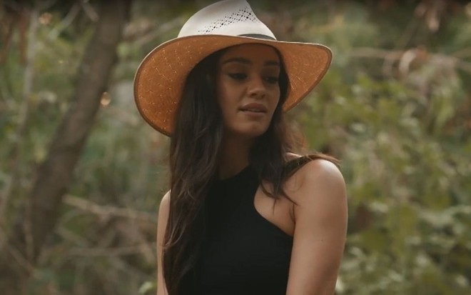 Em cena de Renascer, Sophie Charlotte está falando com alguém; ela usa um chapéu e blusa de alça preta