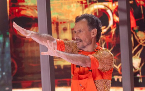 Enrique Diaz usa blusa laranja e está com os braços esticados durante sua apresentação na Dança dos Famosos