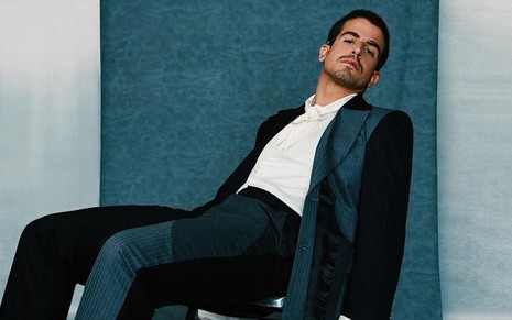 Enzo Celulari usa blusa branca e blazer e calça cinza e preta; ele está encostado em uma cadeira