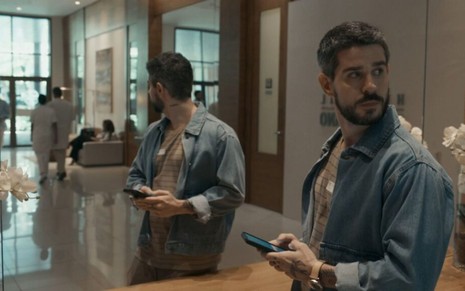 Em cena de Renascer, Pedro Neschling está olhando para o lado, com o celular em mãos