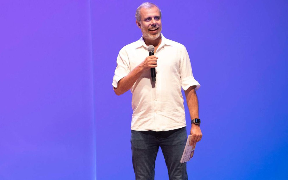 O jornalista Erick Brêtas em evento da Globo; em pé, ele veste camisa branca sobre fundo azul