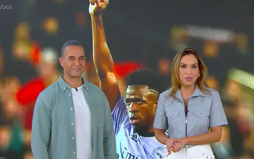 Fabricio Battaglini e Talitha Morete estão em frente a telão com imagem de Vinicius Jr.