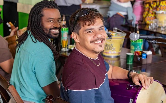 Jão (Fabrício Boliveira) e Sidney (Adanilo) sorriem para a foto; eles estão em mesa de bar com instrumentos musicais