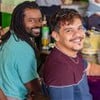 Jão (Fabrício Boliveira) e Sidney (Adanilo) sorriem para a foto; eles estão em mesa de bar com instrumentos musicais