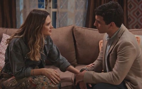 Os atores Juliana Paiva e Henrique Barreira frente a frente, sentados em sofá, olhando um para o outro em cena de Família É Tudo