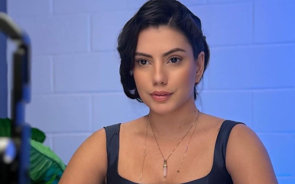 Fernanda Bande usa uma blusa de alça azul; ela encara um ponto fixo, séria