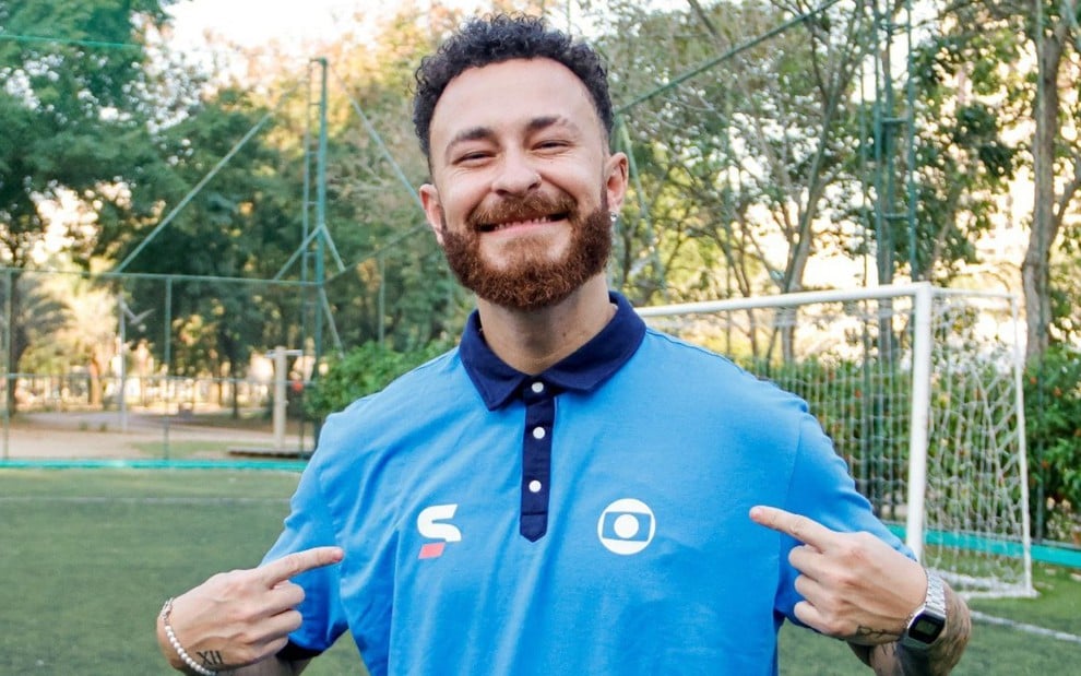 Fred Bruno veste uma camiseta com as logomarcas da Globo e do SporTV e sorri, apontando para os símbolos