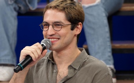 Gabriel Leone está com uma blusa cinza, usa óculos e está falando ao microfone sorrindo no palco do Altas Horas