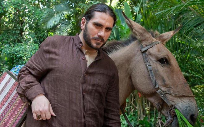 Caracterizado como seu personagem em Renascer, Gabriel Sater usa camisa marrom ao lado de um cavalo