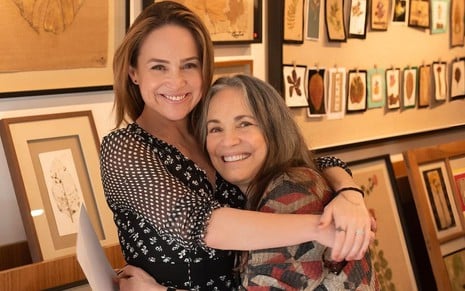 Gabriela Duarte abraça Regina Duarte; as duas estão em uma galeria de arte repleta de quadros expostos