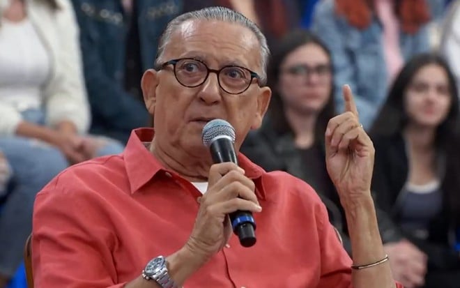 Galvão Bueno usa uma camisa laranja e um óculos de aros pretos. Ele encara Serginho Groisman --fora do quadro-- e segura um microfone. O semblante está sério.