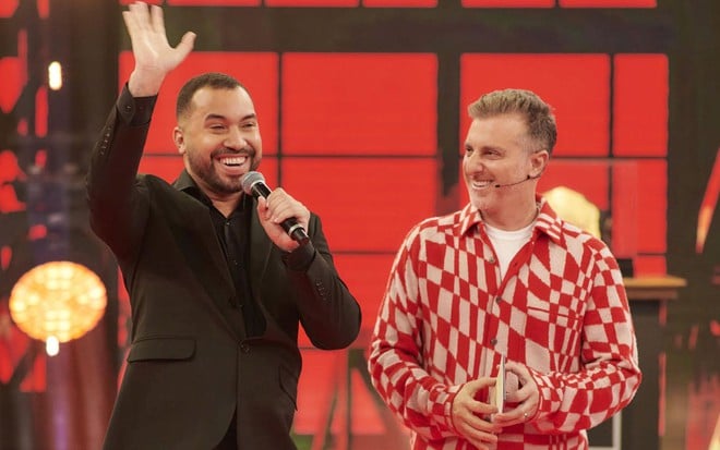 Vestido de preto, Gil do Vigor está ao lado de Luciano Huck no palco do Domingão com Huck