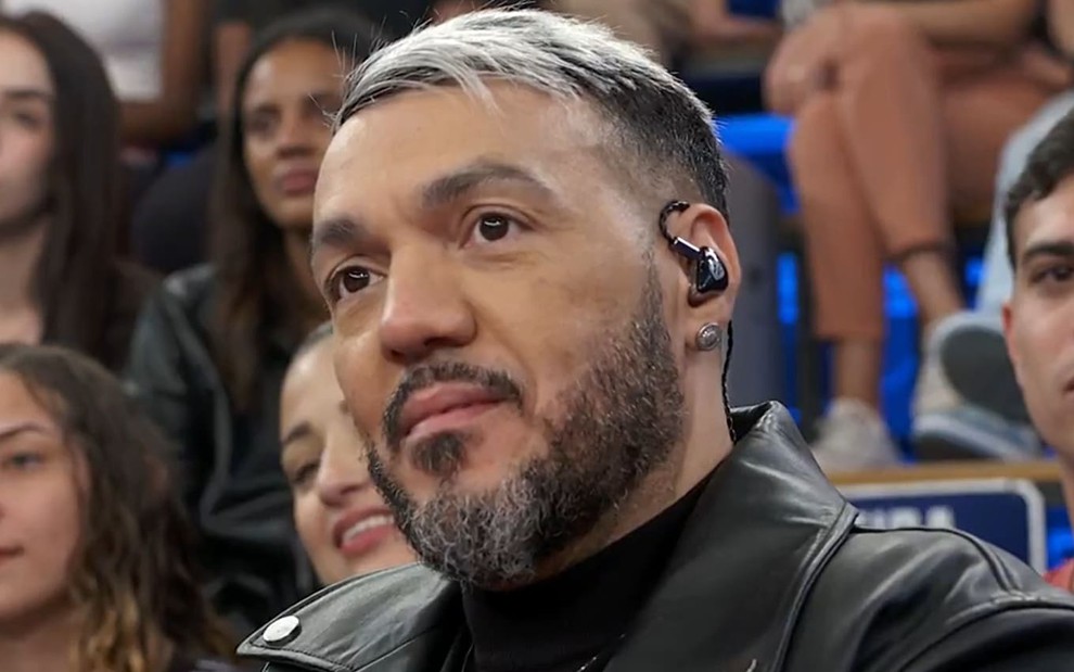 Belo usa uma camiseta preta e uma jaqueta preta de couro; o cabelo está parcialmente platinado, e ele dá um sorriso enquanto segura o microfone