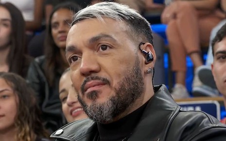 Belo usa uma camiseta preta e uma jaqueta preta de couro; o cabelo está parcialmente platinado, e ele dá um sorriso enquanto segura o microfone