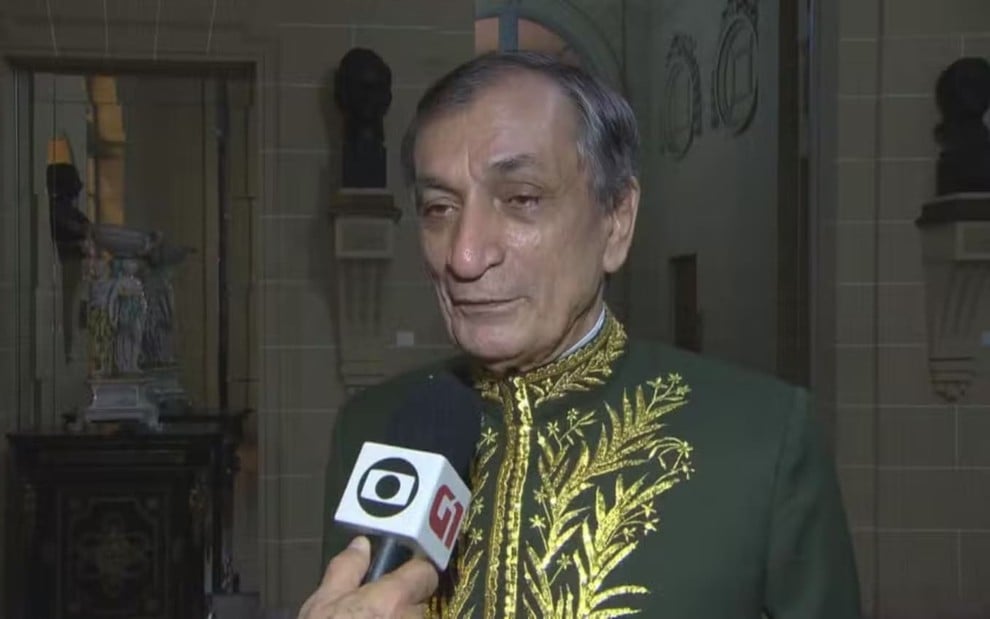 Antonio Cícero usa uma roupa cerimonial, com detalhes em dourado; ele dá entrevista para a Globo