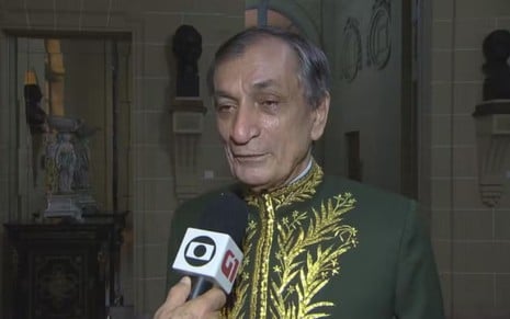 Antonio Cícero usa uma roupa cerimonial, com detalhes em dourado; ele dá entrevista para a Globo