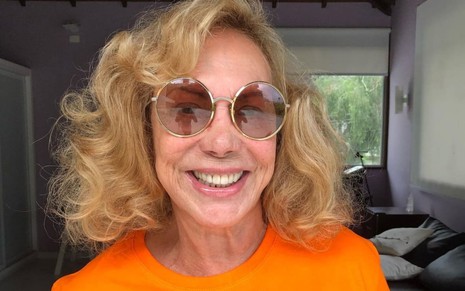 Arlete Salles usa uma blusa laranja e um óculos escuros e sorri para a câmera. O cabelo é loiro e curto.