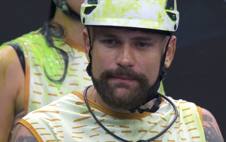 Vinicius Rodrigues usa uma blusa listrada verde e laranja e um capacete amarelado. Ele parece tenso durante prova do BBB 24