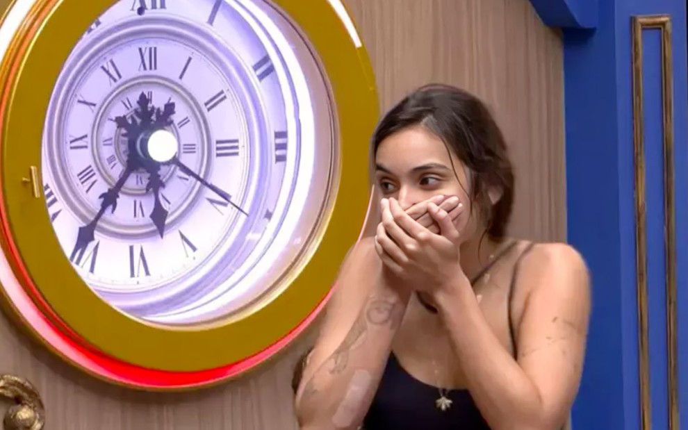 Vanessa Lopes leva a mão à boca em expressão de surpresa ao lado de um relógio com números romanos no BBB 24