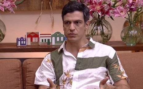 Mateus Solano usa uma camisa listrada branca e verde-musgo; o semblante está sério. O ator está sentado num sofá, na frente de uma série de plantas --o cenário do É de Casa