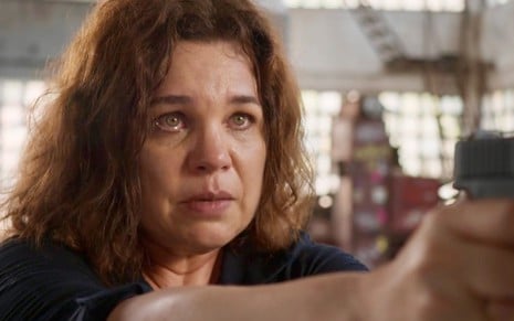 Isabel Teixeira aponta uma arma e está em close como Helena em cena da novela Elas por Elas, da Globo