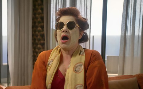 A atriz Isabel Teixeira com uma máscara verde na cara, bobes no cabelo e óculos escuros como Helena em cena de Elas por Elas