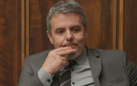 Cassio Gabus Mendes caracterizado como Roberto; ele usa um terno cinza e parece tenso em cena de Elas por Elas
