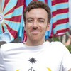 O ator João Côrtes usa camiseta branca e posa caracterizado como Celso em cenário da série Encantado's