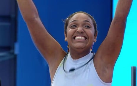 A imagem mostra uma mulher negra sorrindo amplamente, com os braços erguidos em celebração. Ela usa uma blusa sem mangas e está em um ambiente de estúdio com fundo azul.