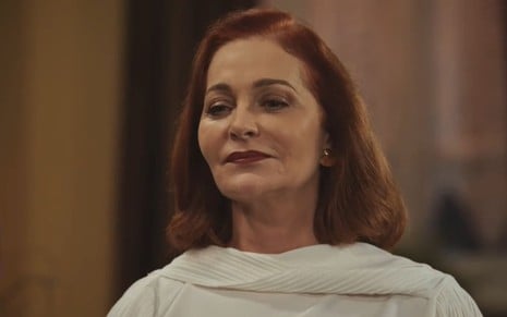 Alexandra Richter caracterizada como Brenda; ela tem os cabelos curtos e ruivos, usa uma blusa branca e está séria em cena de Família É Tudo