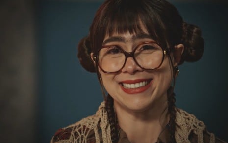 Com monocelha e óculos grandes, Daphne Bozaski sorri com expressão boba em cena de Família É Tudo
