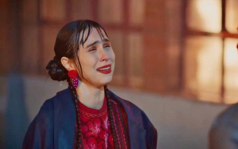 Daphne Bozaski usa blusa vermelha, com casaco azul, e faz cara de choro em cena da novela Família É Tudo, da Globo