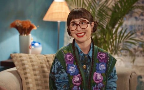 Daphne Boazaski usa roupas coloridas, óculos e tem cabelos presos em cena como Lupita em Família É tudo, da Globo