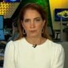 A imagem mostra uma apresentadora de televisão em um estúdio de notícias, com expressão séria, usando um vestido branco e brincos prateados. Ao fundo, vê-se uma redação com monitores e pessoas trabalhando.