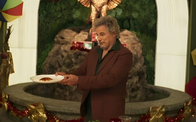 O ator Edson Celulari como Nero em cena de Fuzuê segurando um prato com um pedaço de ave natalina