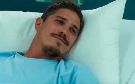Romulo Arantes Neto caracterizado como Julião; ele está deitado num leito de hospital e parece tenso em Fuzuê