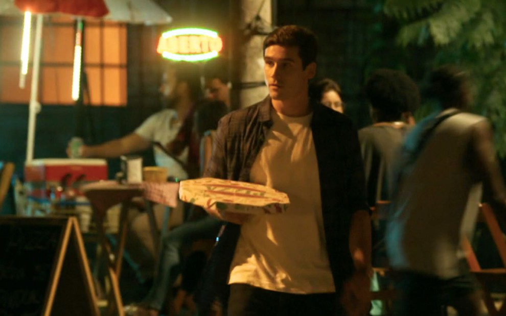 O ator Nicolas Prattes como Miguel carrega uma pizza pequena na mão em cena de Fuzuê