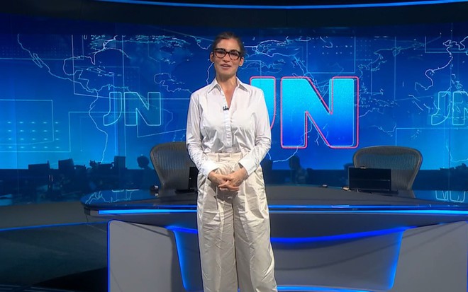 Renata Vasconcellas com uma camisa branca e uma calça bege amarrotadas em frente à bancada do Jornal Nacional