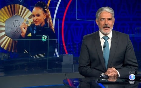 William Bonner está na bancada do Jornal Nacional, com expressão séria; ao fundo, a imagem de Rebeca Andrade com medalha de ouro