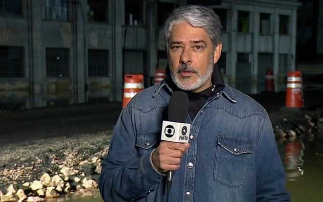 William Bonner com um casaco jeans e um microfone da Globo no Jornal Nacional