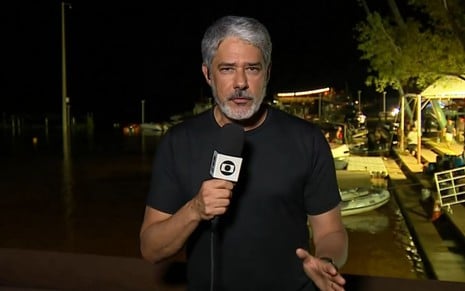 William Bonner com uma camiseta preta apresentando o Jornal Nacional da orla do Guaiba, em Porto Alegre
