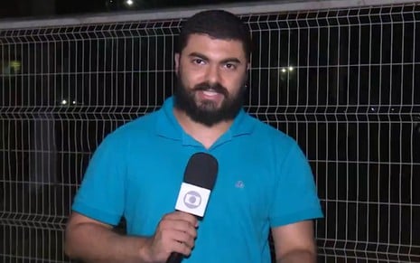 Um homem com uma camisa azul segura um microfone da Globo em frente a um prédio completamente apagado