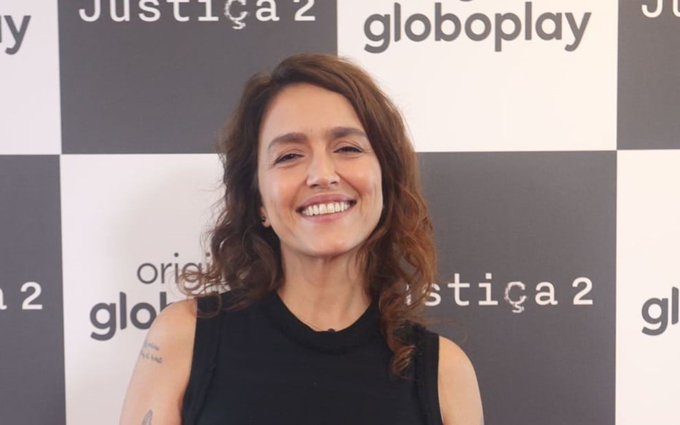 A imagem mostra uma mulher sorridente com cabelos castanhos ondulados, usando uma blusa preta sem mangas. Ao fundo, há um painel com os logos "Globoplay" e "Justiça 2".