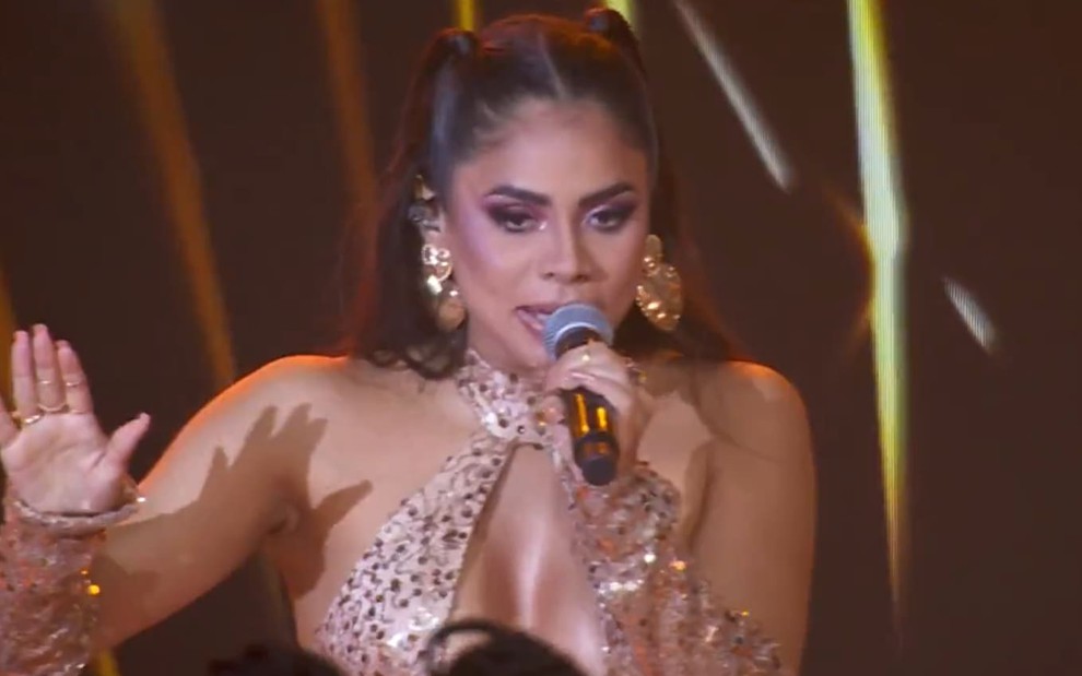 Lexa está com um macacão brilhoso dourado e segura o microfone durante show no BBB 24