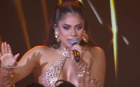 Lexa está com um macacão brilhoso dourado e segura o microfone durante show no BBB 24
