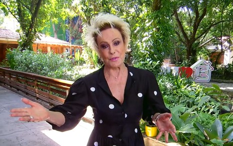 Ana Maria Braga está em frente ao jardim usado agora pelo É de Casa, nos Estúdios Globo; apresentadora está séria e com os braços abertos