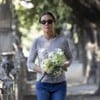 A mulher, de óculos escuros, veste camiseta cinza e calça jeans, caminhando em local arborizado, segurando um buquê de flores brancas e verdes.